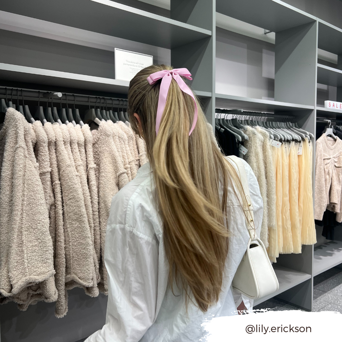 Woman with long hair in a Peony Pink Coconut Lane ribbon bow clip, browsing fuzzy beige jackets in a store.