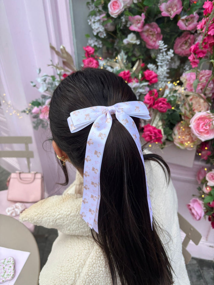Woman with long dark hair wears a beige ribbon bow clip, seated near pink flowers and a light pink purse.