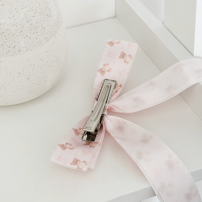 Pink satin ribbon bow clip with teddy bear prints, displayed on a white surface near a vase.