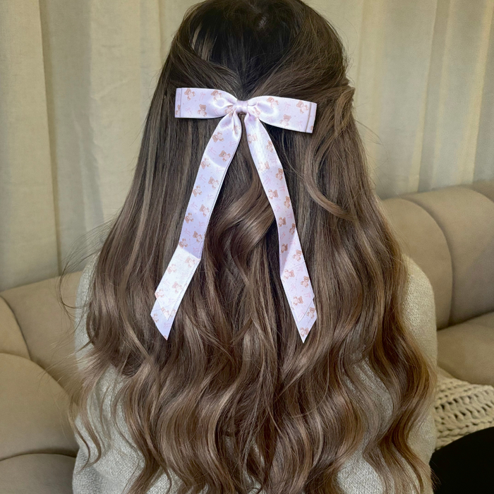 Woman with long wavy brown hair wears a soft beige ribbon bow hair clip, sitting on a beige couch.