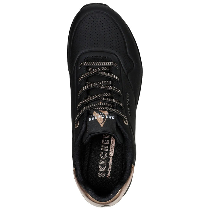 A black sneaker with perforated design and bronze accents sits against a plain background showcasing a sleek silhouette and thick laces highlighting its sporty appearance and cushioning features