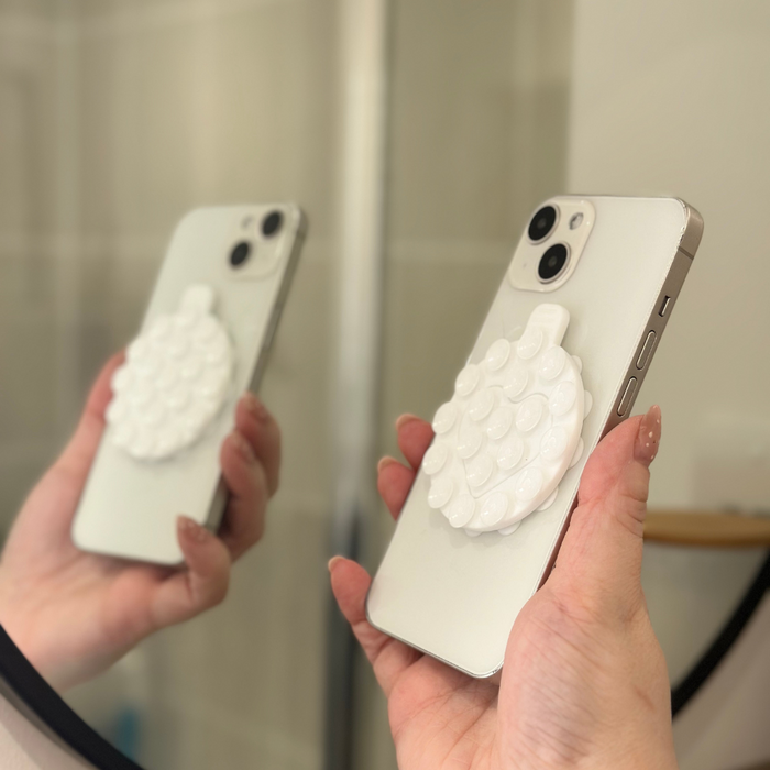 White Coconut Lane suction phone holder secures a phone, both seen in a hand and reflected in a round mirror.