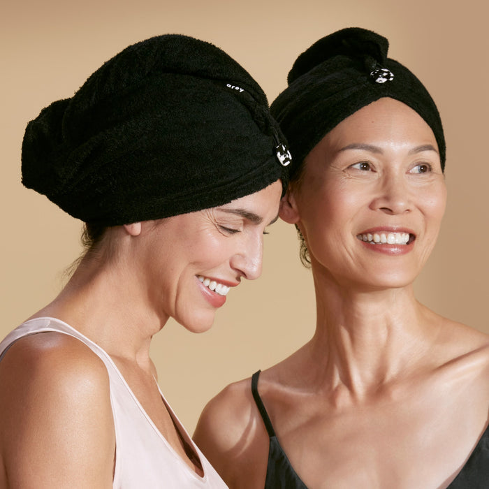Two women smiling, each wearing The Hair Wrap by Arey in black, wrapped around their hair. The background is beige, and both women are dressed in sleeveless tops.