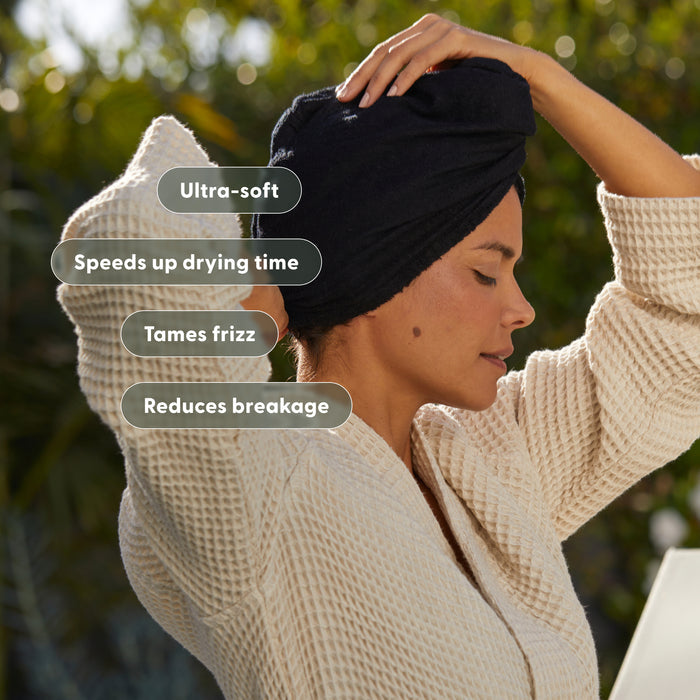 Lifestyle photo of a woman wearing the arey dark hair wrap towel in a turban style, outdoors with a bright bokeh background. Feature badges overlay the image: Ultra-soft, Speeds up drying time, Tames frizz, Reduces breakage.