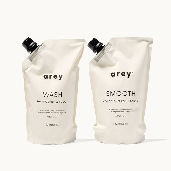Arey Shower Duo Refill Pouches on a cream background. Left: Arey Wash Shampoo Refill Pouch (500ml / 16.9 fl oz) — A gentle cleansing shampoo for stimulating and preserving pigment. All hair types. Right: Arey Smooth Conditioner Refill Pouch (500ml / 16.9 fl oz) — A nourishing conditioner that strengthens and softens. All hair types.