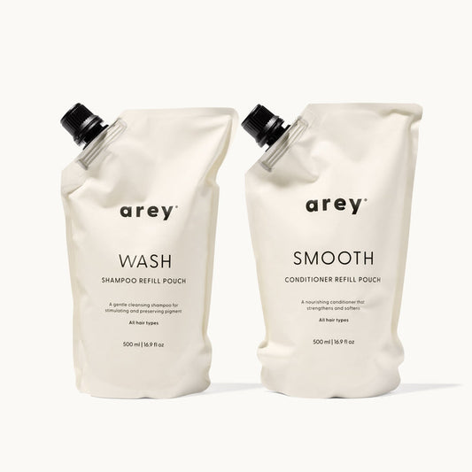 Arey Shower Duo Refill Pouches on a cream background. Left: Arey Wash Shampoo Refill Pouch (500ml / 16.9 fl oz) — A gentle cleansing shampoo for stimulating and preserving pigment. All hair types. Right: Arey Smooth Conditioner Refill Pouch (500ml / 16.9 fl oz) — A nourishing conditioner that strengthens and softens. All hair types.