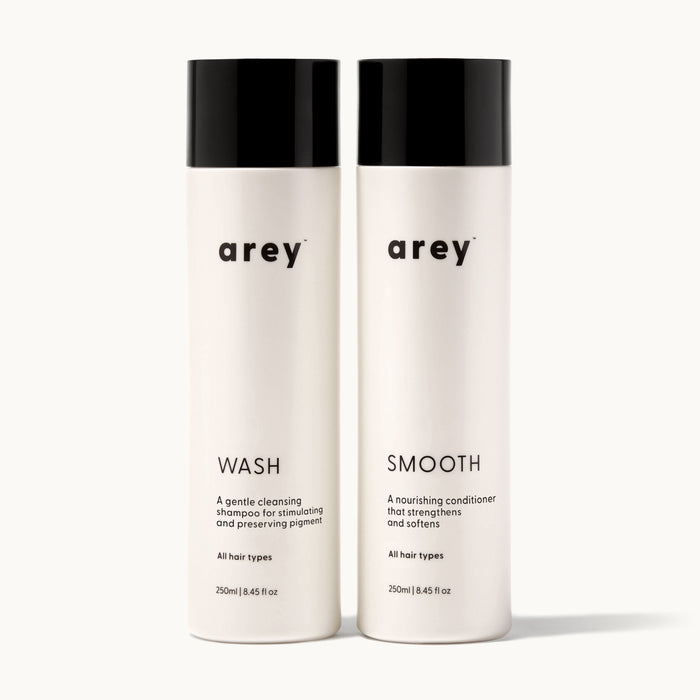 Arey Shower Duo on cream background. Two bottles side by side: Wash (A gentle cleansing shampoo for stimulating and preserving pigment. All hair types. 250ml / 8.45 fl oz) and Smooth (A nourishing conditioner that strengthens and softens. All hair types. 250ml / 8.45 fl oz).