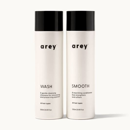 Arey Shower Duo on cream background. Two bottles side by side: Wash (A gentle cleansing shampoo for stimulating and preserving pigment. All hair types. 250ml / 8.45 fl oz) and Smooth (A nourishing conditioner that strengthens and softens. All hair types. 250ml / 8.45 fl oz).
