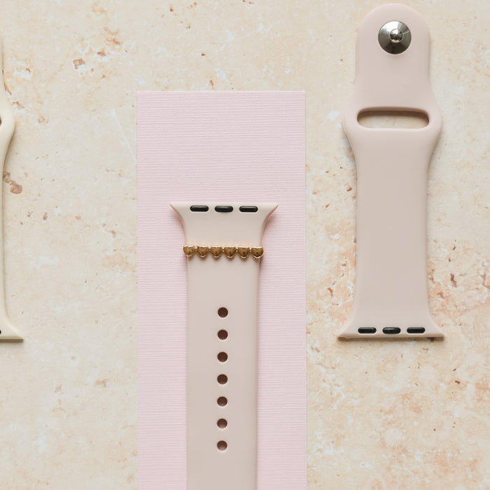 Beige smartwatch bands on marble, one featuring a gold heart charm accessory.
