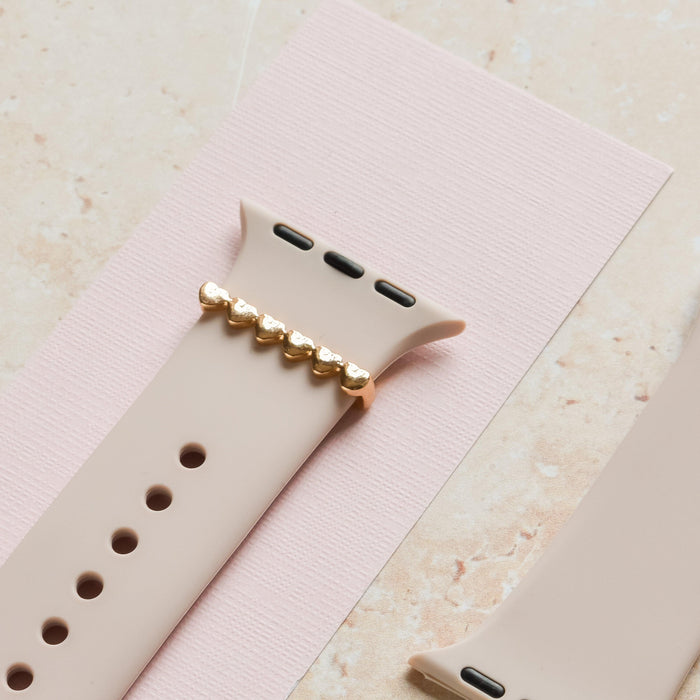 Gold heart watch strap charm from Coconut Lane on a pink textured background with a beige smartwatch band.
