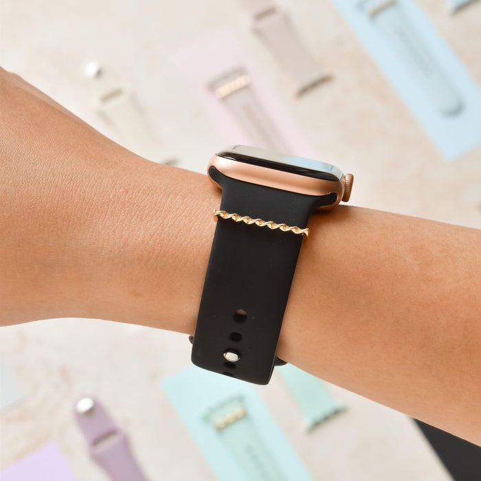 Gold twist watch strap charm by Coconut Lane on a wrist, with blurred colorful watchbands in the background.