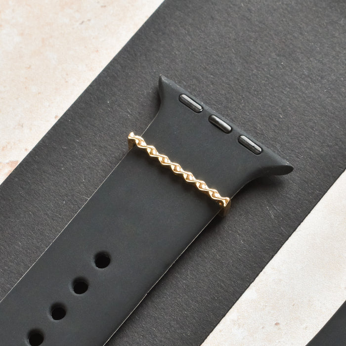 Gold twist watch strap charm by Coconut Lane on a black band, set against a black and beige background.