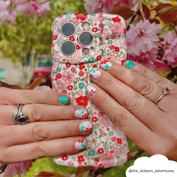 Hands with floral nail art hold a pink flower-patterned wavy phone case next to pink cherry blossoms.