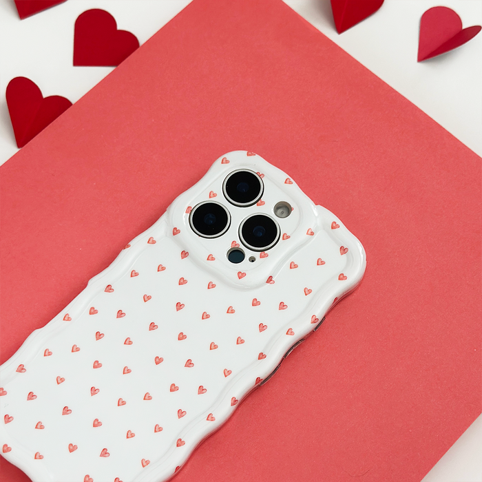 White phone case with wavy edges and mini heart pattern, surrounded by paper hearts on a red surface.