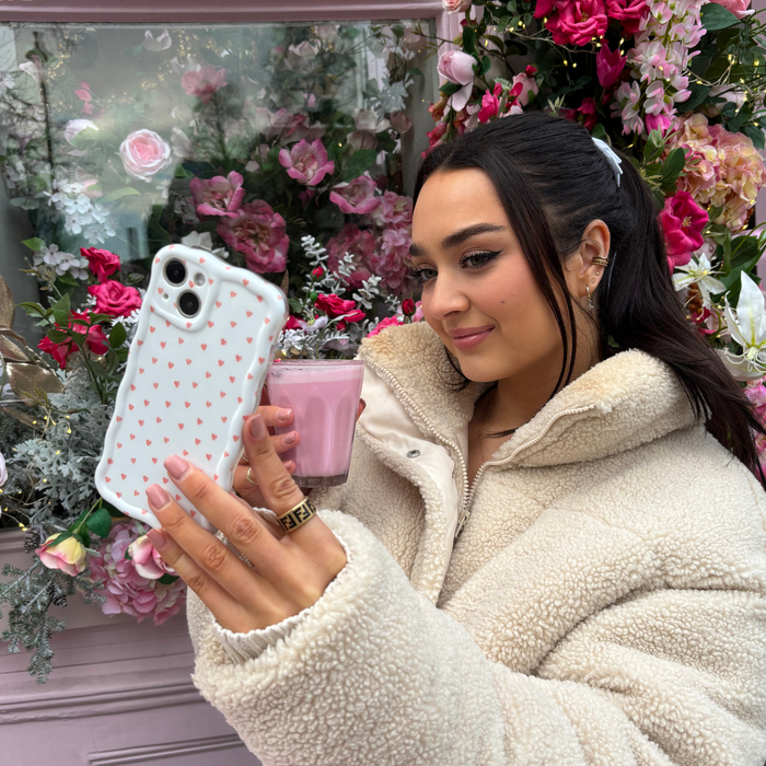 Woman in a fluffy jacket holds a pink drink and takes a selfie with a wavy phone case featuring pink mini hearts.