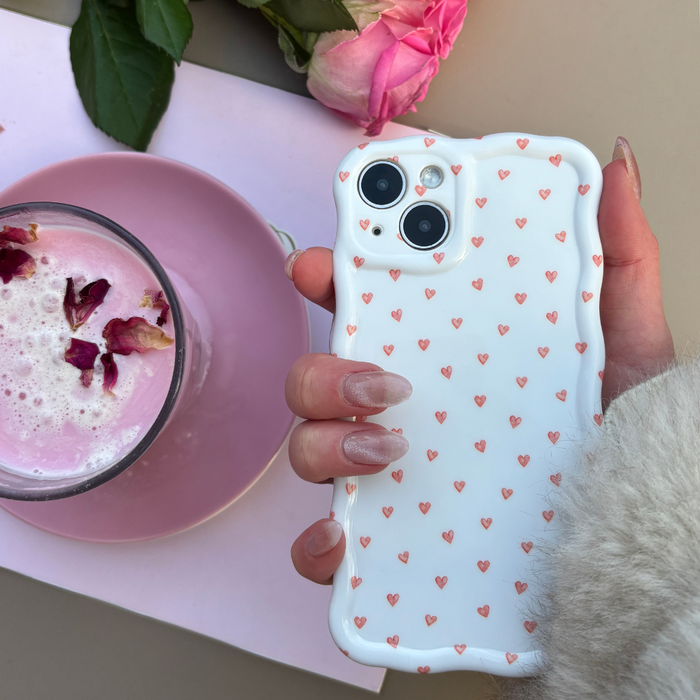 Hand with manicured nails holds a wavy phone case with mini heart pattern beside a pink latte topped with rose petals.