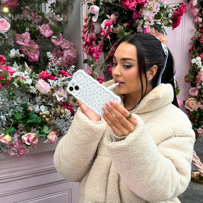 Woman in cream coat taking selfie by pink and white flowers, holding a wavy phone case with mini heart pattern.