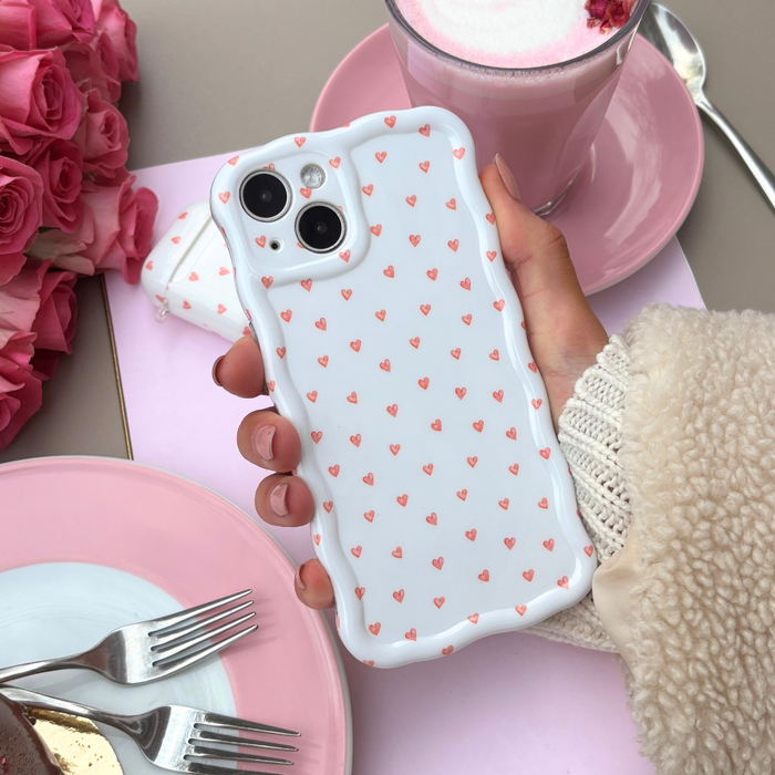 Hand holding Coconut Lane Wavy Phone Case with mini heart pattern, shown beside pink roses and a pink drink on a table.