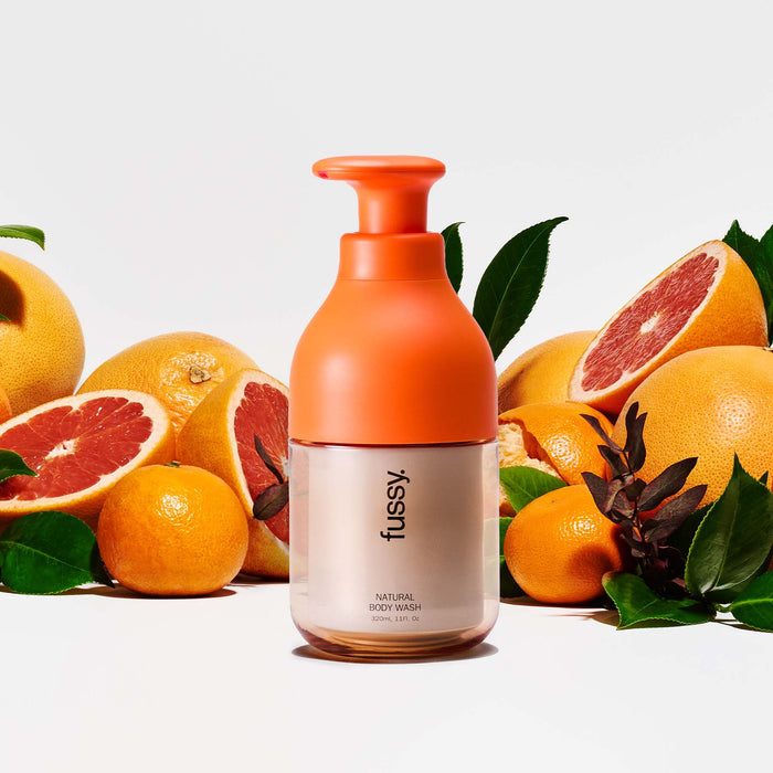 Fussy's natural, refillable Wide Eyed body wash in an Orange pump bottle, set against a background of orange halves and green leaves.